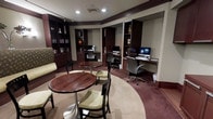 This single-floor space functions as a combined business center and lounge area, thoughtfully designed to accommodate both productivity and relaxation. The room features rich, dark wood built-in shelving units that line the walls, displaying books and decorative accents that add a refined, library-like character to the space. Multiple computer workstations are positioned along the back wall, each equipped with a desktop monitor and an ergonomic office chair, providing a practical and private setting for work or research. A recessed ceiling with recessed lighting creates a warm, focused ambiance throughout the room. At the center, a circular area rug defines a casual seating zone furnished with round pedestal tables and cushioned dining-height chairs in a neutral green upholstery, offering a comfortable spot for informal meetings or leisure. The deep burgundy carpeting that borders the central rug adds a classic, hospitality-inspired aesthetic. The overall design balances a professional atmosphere with welcoming comfort, making the space well-suited as a shared amenity for guests or residents seeking both a functional workspace and a relaxed setting.