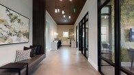 Located at 1701 J N Pease Place in Charlotte, North Carolina, this single-floor amenity space offers a thoughtfully curated environment designed for both productivity and leisure. The interior showcases a contemporary aesthetic defined by large-format porcelain tile flooring, elevated ceilings, and a refined material palette of dark metals, warm wood accents, and crisp white surfaces.

The welcoming entry corridor features an artistic cluster chandelier with exposed Edison-style bulbs set against a dark ceiling, flanked by a built-in upholstered bench adorned with decorative pillows and original abstract artwork. Adjacent to the entry is a dedicated cyber lounge outfitted with multiple desktop workstations beneath minimalist pendant lighting.

Black steel-framed glass partition walls frame a series of private meeting rooms, allowing natural light to flow freely while maintaining acoustic separation. These rooms feature textured accent walls, sophisticated furnishings, and outdoor views.

The expansive clubroom is a social centerpiece, boasting soaring ceilings with clerestory windows, a wall-mounted television, leather seating, and a shuffleboard table. A separate game lounge includes a billiards table, tufted banquette seating, wooden ceiling accents, and high-top bar seating. A wet bar area with glass shelving, a beverage refrigerator, and garden views completes the space.