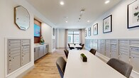 Located in Houston, TX, this well-appointed single-floor community lobby space offers a welcoming and functional environment for residents. The open layout seamlessly integrates a lounge area, mail room, and collaborative seating zones under a clean, modern design aesthetic.

Light wood-look tile flooring runs throughout, lending warmth and continuity to the space. Floor-to-ceiling windows dressed with neutral linen drapes flood the lounge area with natural light and provide views of the surrounding greenery outside. Plush swivel lounge chairs arranged around a low round table create an inviting gathering spot.

The mail area is thoughtfully designed, with rows of sleek, wall-mounted mailboxes lining multiple walls. Framed botanical artwork displayed above the mailboxes adds a refined, decorative touch. A central accent wall, finished in a deep warm tone, houses a mounted flat-screen display and flanking octagonal mirrors above a gray shaker-style console cabinet, serving as a functional and visual focal point.

White conference-style tables with upholstered task chairs provide a practical workspace for residents. Recessed lighting throughout ensures consistent illumination, complemented by ceiling-integrated speakers. The overall ambiance is clean, modern, and inviting — balancing utility with a polished residential aesthetic.