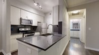 This single-floor residence showcases a contemporary design with a cohesive neutral color palette throughout. The kitchen serves as a central highlight, featuring white cabinetry, dark quartz countertops, stainless steel appliances including a range, microwave, and refrigerator, and a peninsula with an undermount sink — all illuminated by track lighting overhead. Dark hardwood-style flooring flows through the kitchen and into the open living area, which is accented by crown molding and a ceiling fan. A sliding glass door in the living space allows natural light to filter in and provides access to an outdoor area. The bedrooms offer plush carpeting, ceiling fans, and ample natural light from windows, creating comfortable retreats. A hallway bathroom includes a tub-shower combination with tiled surround, a toilet, and a vanity with dark quartz countertops and white cabinetry that mirrors the kitchen's aesthetic. The consistent use of neutral tones, modern finishes, and durable flooring materials throughout lends the space a clean, move-in-ready appeal.
