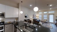 This single-floor residence showcases a thoughtfully designed open-concept layout that seamlessly connects the kitchen and living areas into one cohesive, functional space. The kitchen is outfitted with dark granite countertops, a stainless steel undermount sink with a sleek pull-down faucet, and crisp white cabinetry that adds a clean, modern contrast. A pair of globe pendant lights hang dramatically above the kitchen island, providing both task lighting and a striking visual focal point that carries the eye into the living space. The vaulted ceiling amplifies the sense of openness and airiness throughout. The living area is warmly appointed with a neutral-toned sofa, a bold blue accent chair, a patterned area rug, and curated décor including wall art and a round mirror. Recessed lighting and a floor lamp contribute to a layered, inviting ambiance. Windows dressed with blinds allow natural light to filter in, brightening the space throughout the day. The overall design aesthetic is contemporary and cohesive, balancing clean architectural lines with warm, livable furnishings to create a space that is both visually appealing and highly functional.