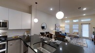 This single-floor residence showcases a thoughtfully designed open-concept layout that seamlessly connects the kitchen and living areas into one cohesive, functional space. The kitchen is outfitted with dark granite countertops, a stainless steel undermount sink with a sleek pull-down faucet, and crisp white cabinetry that adds a clean, modern contrast. A pair of globe pendant lights hang dramatically above the kitchen island, providing both task lighting and a striking visual focal point that carries the eye into the living space. The vaulted ceiling amplifies the sense of openness and airiness throughout. The living area is warmly appointed with a neutral-toned sofa, a bold blue accent chair, a patterned area rug, and curated décor including wall art and a round mirror. Recessed lighting and a floor lamp contribute to a layered, inviting ambiance. Windows dressed with blinds allow natural light to filter in, brightening the space throughout the day. The overall design aesthetic is contemporary and cohesive, balancing clean architectural lines with warm, livable furnishings to create a space that is both visually appealing and highly functional.