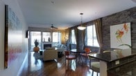 This single-floor residence presents an open-concept layout that seamlessly integrates the living, dining, and kitchen areas into one cohesive, flowing space. Rich hardwood floors run throughout, reflecting the ambient light and lending warmth to the interior. A striking stacked-stone accent wall serves as a defining architectural feature, adding texture and depth to the space. The living area is anchored by a classic fireplace and furnished with contemporary neutral-toned seating, complemented by a ceiling fan overhead. Large windows dressed with floor-length drapes in earthy tones line the walls, allowing generous natural light to fill the room and offering views to the exterior. The dining area accommodates a round table with orange upholstered chairs beneath a modern pendant light fixture, creating an intimate yet stylish setting. Adjacent to the dining space, a glimpse of the kitchen counter is visible, hinting at a well-integrated culinary area. The overall ambiance balances urban sophistication with rustic warmth, achieved through the interplay of stone, wood, and carefully curated furnishings throughout.