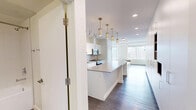 Located at 10045 111 St NW in Edmonton, Alberta, this efficiently designed single-floor unit offers a contemporary open-concept layout that maximizes every square foot. The kitchen is a standout feature, showcasing sleek white cabinetry, a grey tile backsplash, stainless steel appliances including a full-size refrigerator, electric range, and built-in microwave, along with a generous island topped with a light quartz surface — ideal for meal prep or casual dining. Stylish pendant lighting in a brushed gold finish adds a modern touch overhead, while dark hardwood-style flooring runs throughout, grounding the bright, neutral palette. Abundant built-in storage cabinetry lines one wall of the open living area, which also features a built-in low credenza beneath large windows. Those windows frame impressive views of Edmonton's downtown skyline, flooding the space with natural light. The bathroom is clean and minimal, finished with large-format white wall tiles, a soaker tub with overhead shower, glass shelving, and grey floor tile. Overall, the unit presents a cohesive modern aesthetic with high-quality finishes throughout.
