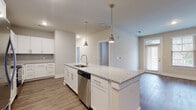 This one-bedroom, one-bathroom residence at Annabelle on Main in Duluth, Georgia offers a thoughtfully designed single-floor layout with contemporary finishes throughout. The open-concept kitchen serves as the centerpiece of the living area, anchored by a large island topped with light granite countertops and fitted with an undermount sink and stainless steel dishwasher. Pendant lights with brushed nickel fixtures hang above the island, adding an elegant touch. The kitchen is further equipped with white shaker-style cabinetry extending to the ceiling, a subway tile backsplash, and a full suite of stainless steel appliances including a refrigerator, electric range, and over-the-range microwave. Wood-look plank flooring runs throughout the main living areas, complementing the neutral gray walls and clean white trim. The living area is generously proportioned, offering flexible arrangement possibilities. The bedroom features plush carpet flooring, a ceiling fan, and a window that invites natural light. The bathroom includes a glass-enclosed walk-in shower and wood-look tile flooring, maintaining the cohesive aesthetic found throughout the residence.