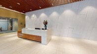 Located at 710 12th St in Arlington, Virginia, this polished commercial lobby presents a sophisticated first impression through a carefully curated blend of materials and contemporary design. The entry level is anchored by a striking reception desk finished in crisp white stone and warm wood accents, set against a dramatic textured marble wall illuminated by recessed lighting above. A signature slatted wood ceiling runs throughout, adding warmth and architectural depth to the space.

The expansive lobby floor is laid with large-format cream tiles that reflect light and contribute to an open, airy atmosphere. A large-scale map mural adorns one wall, offering a local geographic reference and a distinctive design focal point. Elevator bays, framed by rich wood paneling and clean marble surfaces, provide seamless vertical access throughout the building.

A dedicated lounge area transitions the space into a comfortable gathering zone, furnished with mid-century modern swivel chairs in bold blue and red tones, complemented by round wood coffee tables and ambient floor lamps. A dramatic circular chandelier crowns this seating area, while floor-to-ceiling sheer curtains filter abundant natural light and frame views of the surrounding Arlington streetscape. Glass-partitioned zones allow for versatile use of the space while maintaining visual connectivity throughout.