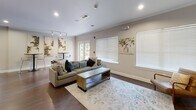Located at 2001 Dogwood Dr in Fredericksburg, Virginia, this single-floor community clubhouse offers a thoughtfully designed gathering space with a variety of distinct areas tailored for socializing and recreation. The grand main lounge commands immediate attention with soaring double-height ceilings, a striking stone fireplace, large clerestory windows that flood the room with natural light, and rich hardwood flooring throughout. A ceiling fan anchors the lofty space, which is furnished with comfortable mid-century modern seating arrangements. Adjacent to the lounge, a spacious full kitchen features dark espresso cabinetry, granite countertops, a mosaic tile backsplash, and a built-in wine rack. A broad granite-topped island with bar seating connects the kitchen to a lounge area outfitted with built-in shelving and a mounted television. A casual game and dining area includes a round table with lounge seating, a billiards table, and additional cabinetry with counter space. A separate sitting room offers plush sectional seating, a game table, and French doors opening directly to a lush outdoor patio. High-top café tables set against decorative wall art provide an intimate nook for smaller gatherings. Throughout, hardwood floors, recessed lighting, and generous window treatments create a cohesive, inviting ambiance.