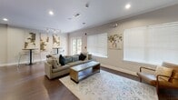 Located at 2001 Dogwood Dr in Fredericksburg, Virginia, this single-floor community clubhouse offers a thoughtfully designed gathering space with a variety of distinct areas tailored for socializing and recreation. The grand main lounge commands immediate attention with soaring double-height ceilings, a striking stone fireplace, large clerestory windows that flood the room with natural light, and rich hardwood flooring throughout. A ceiling fan anchors the lofty space, which is furnished with comfortable mid-century modern seating arrangements. Adjacent to the lounge, a spacious full kitchen features dark espresso cabinetry, granite countertops, a mosaic tile backsplash, and a built-in wine rack. A broad granite-topped island with bar seating connects the kitchen to a lounge area outfitted with built-in shelving and a mounted television. A casual game and dining area includes a round table with lounge seating, a billiards table, and additional cabinetry with counter space. A separate sitting room offers plush sectional seating, a game table, and French doors opening directly to a lush outdoor patio. High-top café tables set against decorative wall art provide an intimate nook for smaller gatherings. Throughout, hardwood floors, recessed lighting, and generous window treatments create a cohesive, inviting ambiance.