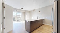 This single-floor residence presents a contemporary open-concept layout with well-defined living spaces and consistent modern finishes throughout. The kitchen serves as a central feature, anchoring the living area with a substantial island finished in dark wood cabinetry and a crisp white countertop. Stainless steel appliances, including a refrigerator, microwave, and range, are complemented by white upper cabinetry, a subway tile backsplash, and pendant lighting overhead. A stainless steel sink is integrated into the island, which also provides bar-style seating. Light hardwood flooring extends throughout the main living areas, contributing to a bright and cohesive aesthetic. Large sliding glass doors and oversized windows invite abundant natural light and provide elevated views of the surrounding urban cityscape. The bedrooms are finished with neutral-toned carpet, large windows framing city views, and glass-panel doors that add a modern touch. The bathroom is well-appointed with a glass-enclosed walk-in shower clad in neutral tile, a dark wood vanity with an integrated sink, a framed mirror, and hardwood flooring, completing the space with a clean, contemporary feel.