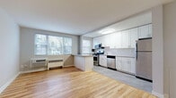 This single-floor unit is finished with light hardwood flooring that runs consistently throughout the living areas and bedrooms, creating a warm, cohesive aesthetic. The open-concept living and kitchen area is bright and airy, benefiting from a generous bank of windows that invite natural light and offer views of surrounding greenery. A peninsula counter separates the kitchen from the living space, providing additional workspace and a natural division between the two areas.

The kitchen is well-appointed with white shaker-style cabinetry extending to the ceiling, offering abundant storage. Granite-style countertops complement the subway tile backsplash, while a full suite of stainless steel appliances — including a gas range, over-the-range microwave, dishwasher, and refrigerator — ensures modern functionality. The flooring transitions to a neutral tile in the kitchen area.

The bedrooms are comfortably sized with neutral gray walls, hardwood floors, and individual windows that provide natural light. One bedroom features a ceiling fan, while both rooms include wall-mounted heating units. An entry area includes access to in-unit laundry. The bathroom features a full soaking tub with white square tile surround, chrome fixtures, and clean white finishes throughout.