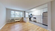 This single-floor unit is finished with light hardwood flooring that runs consistently throughout the living areas and bedrooms, creating a warm, cohesive aesthetic. The open-concept living and kitchen area is bright and airy, benefiting from a generous bank of windows that invite natural light and offer views of surrounding greenery. A peninsula counter separates the kitchen from the living space, providing additional workspace and a natural division between the two areas.

The kitchen is well-appointed with white shaker-style cabinetry extending to the ceiling, offering abundant storage. Granite-style countertops complement the subway tile backsplash, while a full suite of stainless steel appliances — including a gas range, over-the-range microwave, dishwasher, and refrigerator — ensures modern functionality. The flooring transitions to a neutral tile in the kitchen area.

The bedrooms are comfortably sized with neutral gray walls, hardwood floors, and individual windows that provide natural light. One bedroom features a ceiling fan, while both rooms include wall-mounted heating units. An entry area includes access to in-unit laundry. The bathroom features a full soaking tub with white square tile surround, chrome fixtures, and clean white finishes throughout.