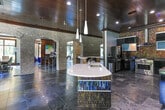 Located at 100 Perimeter Lofts Circle in Dunwoody, Georgia, this single-floor leasing and amenity space makes a strong architectural impression through its thoughtful blend of industrial and refined design elements. Expansive dark slate tile flooring runs throughout the open layout, contrasted beautifully by warm reclaimed wood paneling and rich wood-planked ceilings with decorative crown molding. Signature arched doorways framed in light natural brick create a series of connected zones, lending the space both visual depth and a sense of grand scale. A prominent reclaimed wood accent wall serves as the focal point of the lounge area, anchoring a wall-mounted flat-screen display flanked by tufted deep-blue velvet chairs and a vibrant multicolored area rug. Large floor-to-ceiling windows flood the space with natural light and offer views of the landscaped exterior. A workstation area equipped with iMac computers and adjustable bar-height seating supports leasing operations, while additional rooms provide dedicated office and meeting spaces. A stylish reception bar with illuminated blue tile detailing adds a contemporary accent. Throughout the space, the interplay of exposed brick, polished surfaces, and curated furnishings establishes a sophisticated, welcoming ambiance suited to a premier residential community.