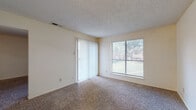 This single-floor residential unit offers a practical and comfortable layout designed for everyday living. The living area is generously sized, featuring wall-to-wall carpet and a textured ceiling that adds subtle dimension to the space. Large windows fitted with blinds allow abundant natural light to fill the room, while a sliding glass door with vertical blinds provides outdoor access and adds to the bright, open feel of the space. The bedroom is similarly carpeted and well-lit, with a window overlooking greenery outside, creating a calm and restful environment. A wide doorway connects the bedroom to the adjoining living space, maintaining an open flow throughout. The kitchen is equipped with warm wood-toned cabinetry — both upper and lower — offering ample storage. It features light-toned countertops, a stainless steel sink, an electric range with overhead ventilation, and wood-look flooring that adds warmth and is easy to maintain. The bathroom continues the wood-look flooring and includes a full-size soaking tub with white tile surround, a toilet, and a vanity with rich wood cabinetry, light countertops, and a wide mirror. Overall, the unit presents a clean, functional living environment with consistent finishes throughout.