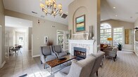 Located at 619 Park Hill Dr in Fairlawn, Ohio, this single-floor community space offers an elegant and inviting atmosphere defined by dramatic vaulted ceilings and thoughtful architectural detailing. The living area serves as the heart of the space, centered around a stunning white-mantled fireplace adorned with hexagonal tile surround and a graceful arched niche above. Warm herringbone-patterned hardwood flooring anchors the seating area, complemented by neutral-toned walls that create a calm, sophisticated ambiance. A classic chandelier overhead adds a refined finishing touch. Large windows throughout the space flood the interior with abundant natural light and provide pleasant views of the surrounding outdoor greenery, enhancing the open, airy feel. The dining area features a round wooden table set beside floor-to-ceiling windows that further emphasize the connection to the outdoors. A dedicated display wall with a mounted flat-screen television and informational panels offers a functional focal point near the entry, which is accessed through a glass-paneled door. The kitchen is outfitted with rich dark cabinetry, granite countertops, a stainless steel refrigerator, and tile flooring, providing a practical and well-appointed space for food preparation and service. Together, these areas combine comfort, functionality, and classic design.