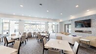 Located at 12501 Ridgedale Dr in Minnetonka, Minnesota, this single-floor amenity space is designed with a clean, contemporary aesthetic throughout. The open-concept layout seamlessly connects a fully equipped kitchen, a dining area, and a comfortable lounge, making it well-suited for gatherings and community events.

The kitchen is outfitted with stainless steel appliances, including a gas range with a stainless steel hood, double wall ovens, a full-size refrigerator, and a microwave. Light wood cabinetry and a large white quartz island with bar seating add both functionality and a polished, modern finish. Light hardwood flooring grounds the kitchen area.

The dining section features multiple white-topped tables with sleek upholstered chairs arranged over patterned carpet, accommodating a generous number of guests. Floor-to-ceiling windows along one wall flood the area with natural light and offer views of an outdoor patio and pergola structure beyond.

The lounge area serves as a striking focal point, anchored by a floor-to-ceiling stacked stone accent wall housing a linear gas fireplace below a wall-mounted flat-screen TV. Cream-toned upholstered sofas and armchairs are arranged around a low white coffee table, creating an inviting and relaxed seating area. Framed glass double doors on either side of the fireplace wall lead to additional rooms, and vibrant artwork adds a curated touch to the space.