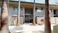 Located at 3131 Cauby St in San Diego, this courtyard offers a well-maintained outdoor communal space that embodies classic Southern California residential design. The centerpiece of the courtyard is a wide, concrete walkway that runs the length of the property, flanked on both sides by tall, mature palm trees that lend a distinctly tropical character to the environment. The ground-level landscaping features a clean gravel border with low-maintenance desert plants and accents of vibrant red foliage, creating a visually appealing contrast against the neutral-toned stucco building exteriors. Privacy fencing in a warm taupe finish lines portions of the walkway, providing defined personal outdoor spaces for ground-floor residents. Exterior wall-mounted lighting adds functional illumination to the pathways and entryways. A metal staircase with sage-green railings connects the ground level to the upper floor, featuring a practical and sturdy design consistent throughout the complex. The two-story building facades are finished in a smooth stucco with louvered shutters framing the windows, complementing the low-key, relaxed aesthetic of the complex. Overall, the courtyard creates a tranquil, semi-private outdoor corridor that reflects the sunny, laid-back character of San Diego living.
