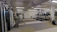 This fitness center offers a practical and organized workout environment spread across a single floor. The space features exposed ductwork and overhead fluorescent lighting mounted to a white ceiling, giving the room an industrial yet clean aesthetic. The flooring consists of a combination of light and grey toned gym-grade material, providing durability and comfort underfoot. Along one side of the room, a row of treadmills equipped with individual digital screens is neatly arranged, allowing users to track their workout metrics. On the opposite side, a multi-functional strength training station is positioned alongside a medicine ball rack stocked with various weighted balls, catering to both resistance and functional training needs. A large mirror along the wall enhances the sense of space and allows users to monitor their form during exercise. The walls are painted white, contributing to a bright and open atmosphere. A waste bin and a utility door are also present, adding to the room's functionality. Overall, the space is efficiently arranged to accommodate a variety of fitness activities in a straightforward, no-frills environment.