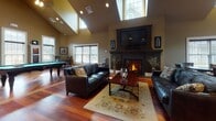 Located at 1085 Hartford Tpke in Vernon Rockville, Connecticut, this single-floor community space blends craftsman-inspired architectural details with modern amenities throughout its generously sized layout. Rich, warm hardwood flooring flows seamlessly from room to room, complementing the dark wood trim, wainscoting, and panel millwork that define the space's refined character.

The main gathering area commands attention with soaring vaulted ceilings and abundant natural light streaming through multiple windows. A dramatic fireplace serves as the focal point, topped by a wall-mounted television and flanked by leather sofas, while a regulation billiards table provides additional entertainment. Recessed lighting and ceiling fans complete the inviting atmosphere.

The kitchen is outfitted with professional-grade stainless steel appliances, including a commercial-range oven with a statement hood, mosaic tile backsplash, and warm wood cabinetry paired with dark stone countertops. A pass-through window connects the kitchen to adjacent areas, enhancing the space's open, social flow.

A dedicated dining lounge area features two-toned walls in soft blue and charcoal with high ceilings, small café-style tables, and a built-in media cabinet with display shelving. The entry foyer showcases bold craftsman double doors, detailed door surrounds, and transom windows, setting an elegant and welcoming tone from arrival.