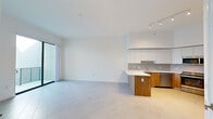 Located at 999 NW Seventh St in Miami, FL, this modern one-bedroom, one-bathroom unit offers a clean, contemporary design throughout its efficiently laid-out single floor. Large-format tile flooring runs consistently across the living areas, reinforcing the bright, minimalist aesthetic defined by crisp white walls and abundant natural light.

The open-concept living and kitchen area forms the heart of the unit. The kitchen is well-appointed with a two-tone design combining white upper cabinetry and warm wood-finish lower cabinets, complemented by a neutral tile backsplash and white quartz countertops. A peninsula with seating overhang provides additional workspace and casual dining. Stainless steel appliances — including a refrigerator, range, microwave, and dishwasher — complete the functional layout. Track lighting overhead highlights the notably high ceilings that lend an airy, expansive feel to the space.

The bedroom features large floor-to-ceiling sliding glass doors that open directly to a private balcony with perforated metal railings, offering views of the surrounding Miami cityscape and filling the room with natural light. The bathroom is sleekly finished with a wood-accented vanity, white quartz countertop, under-mount sink, and a glass-enclosed walk-in shower with white tile surround. A dedicated closet and in-unit storage complete this well-designed urban residence.