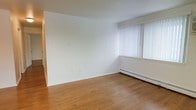 This single-floor space offers a straightforward and functional layout designed for comfortable everyday living. The main living area is finished with warm hardwood flooring that adds a natural tone to the otherwise neutral interior. Bright white walls create a clean, open atmosphere, while a large window dressed with vertical blinds allows ample natural light to fill the room and can be adjusted for privacy. A baseboard heating unit runs along the exterior wall beneath the window, providing efficient climate control throughout the space. The minimalist design of the interior makes it highly adaptable, allowing the space to be configured to suit a variety of needs and furnishing styles. The absence of visual clutter and the use of light tones throughout contribute to an airy, welcoming ambiance. The overall condition of the space appears well-maintained, with clean walls and floors that are move-in ready. This space is well-suited for those seeking a clean, simple environment with practical features and good natural light.