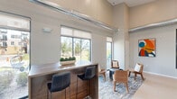 Located at 417 Karen Drive in Woodbury, Minnesota, this single-floor living room serves as a stylish and welcoming communal space. The interior immediately impresses with its soaring ceilings and exposed industrial-style ductwork, lending a contemporary loft aesthetic to the open floor plan. Large, floor-to-ceiling windows and sliding glass doors flood the space with abundant natural light while offering views of the surrounding landscape and neighboring grounds. A warm neutral color palette of soft grays and beiges lines the walls, complemented by light flooring that enhances the sense of openness. The seating area features modern lounge chairs in warm cognac tones arranged around a bold cylindrical coffee table, all anchored by a richly textured, multi-toned area rug that adds visual depth. A wooden bar-height counter with sleek dark stools serves as a functional gathering point, ideal for casual conversation or light work. Curated wall art, including vibrant abstract pieces and geometric prints, adds personality and a creative touch throughout. A striking yellow tile accent wall near the entry creates a bold focal point, while black-framed glass doors and windows reinforce the modern industrial design language. The overall ambiance is inviting, contemporary, and thoughtfully designed for community engagement.