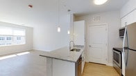 This 1-bedroom, 1-bathroom residence is designed with a clean, modern aesthetic that emphasizes open living and functional design. The space is laid out on a single floor, offering a seamless flow between the kitchen and living areas.

The kitchen serves as a focal point, anchored by a substantial island topped with light gray granite countertops and an undermount stainless steel sink. White cabinetry provides ample storage, while stainless steel appliances add a polished, contemporary touch. Pendant lighting hangs above the island, providing both task lighting and a stylish design element that draws the eye upward toward the bright white ceilings.

Light-toned hardwood flooring runs throughout the space, contributing to the airy, open feel of the interior. The open-concept design connects the kitchen directly to the living area, making the overall layout feel spacious and versatile. Crisp white walls and an abundance of natural light further enhance the bright, welcoming ambiance of the home.

The entry door is conveniently positioned adjacent to the kitchen, providing easy access. Overall, the space strikes a balance between modern style and everyday practicality, making it a comfortable and well-appointed living environment.