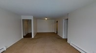 This single-floor residence offers a straightforward and functional layout designed for comfortable everyday living. The main living area is expansive and open, finished with wall-to-wall neutral beige carpeting that extends throughout the space, providing a warm and cohesive feel underfoot. The walls are painted a clean, bright white, creating a light and airy atmosphere that maximizes the sense of space.

The living area features a central flush-mount ceiling light fixture that provides general illumination throughout the room. White painted trim and baseboards line the perimeter, adding a classic, clean finish to the space. A baseboard heating unit is visible along one wall, indicating a reliable heating system integrated into the home.

Multiple doorways branch off from the main living area, connecting to hallways and additional rooms. One opening leads to a corridor with hardwood or dark flooring, suggesting a transition to a distinct area of the home, such as bedrooms or utility spaces. Another doorway opens to a separate hallway with additional doors, hinting at further private rooms beyond. The layout allows for easy circulation and clear separation between the common living space and the more private areas of the residence.

Overall, the space presents a clean, move-in-ready environment with a neutral palette well-suited for a variety of interior design preferences.
