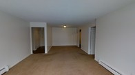 This single-floor residence offers a straightforward and functional layout designed for comfortable everyday living. The main living area is expansive and open, finished with wall-to-wall neutral beige carpeting that extends throughout the space, providing a warm and cohesive feel underfoot. The walls are painted a clean, bright white, creating a light and airy atmosphere that maximizes the sense of space.

The living area features a central flush-mount ceiling light fixture that provides general illumination throughout the room. White painted trim and baseboards line the perimeter, adding a classic, clean finish to the space. A baseboard heating unit is visible along one wall, indicating a reliable heating system integrated into the home.

Multiple doorways branch off from the main living area, connecting to hallways and additional rooms. One opening leads to a corridor with hardwood or dark flooring, suggesting a transition to a distinct area of the home, such as bedrooms or utility spaces. Another doorway opens to a separate hallway with additional doors, hinting at further private rooms beyond. The layout allows for easy circulation and clear separation between the common living space and the more private areas of the residence.

Overall, the space presents a clean, move-in-ready environment with a neutral palette well-suited for a variety of interior design preferences.