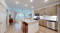 This thoughtfully designed single-floor residence offers a cohesive open-concept layout that maximizes every square foot. The kitchen serves as the centerpiece of the living space, anchored by a granite-topped island with bar seating — ideal for casual dining or entertaining. Warm-toned flat-front cabinetry, a white tile backsplash, and a full suite of stainless steel appliances including a refrigerator, range, microwave, and dishwasher complete the well-appointed kitchen. Light wood-style flooring flows seamlessly from the kitchen into the living room, which is brightened by multiple windows dressed with blinds and grommet curtains, and finished with a ceiling fan overhead. The bedroom offers a comfortable retreat with a stylish barn-style sliding door leading to a dedicated closet. Two bathrooms serve the unit; one features a soaking tub surround finished in large-format marble-look tile with brushed gold fixtures, paired with a wood-toned vanity and granite countertop. Clean white walls and recessed lighting throughout maintain a bright, modern aesthetic, while the consistent use of granite surfaces and warm wood tones creates a unified and contemporary design sensibility across the entire residence.