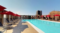 This rooftop amenity space offers an impressive outdoor retreat elevated high above the surrounding cityscape. At its centerpiece sits a generously sized swimming pool with crystal-clear turquoise water, framed by a broad, light-toned concrete sun deck that provides ample room for relaxation and movement. Rows of sling-style lounge chairs line both sides of the pool, each accompanied by bold red market umbrellas that offer shade throughout the day. A covered pergola structure runs along one side of the deck, creating a shaded corridor with additional seating for guests who prefer to stay out of direct sun. At the perimeter of the rooftop, open-air terrace areas extend the usable space further, furnished with bistro-style tables and chairs alongside additional lounge seating, all positioned to take full advantage of the expansive panoramic views. The surrounding cityscape is visible in every direction, with high-rise buildings and the broader urban horizon forming a dynamic backdrop. The open-sky exposure ensures abundant natural light throughout the day, while the elevated position creates a sense of openness and privacy above the city. The overall ambiance is resort-like yet distinctly urban, making this rooftop deck a compelling and functional outdoor amenity.