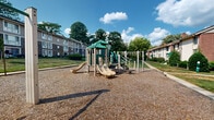 Located in Columbia, Maryland, this outdoor community playground and green space offers a welcoming recreational environment for residents. At its center sits a multi-component play structure featuring a green-roofed platform, dual slides, climbing elements, and overhead monkey bars, all set on a generous wood chip safety surface that cushions the play area. Adjacent seating benches provide a comfortable vantage point for supervising children or simply enjoying the outdoors.

The surrounding landscape is characterized by expansive, well-maintained lawns that roll gently across the grounds, with notable grassy mounds adding natural topographic interest. Mature deciduous trees are distributed throughout the space, offering ample shade and a park-like atmosphere. A network of concrete sidewalks connects various parts of the grounds, providing clear and accessible pathways for pedestrians throughout the community.

Multi-story brick and siding residential buildings frame the perimeter of the playground on multiple sides, creating an enclosed, community-centered feel. Manicured shrubs and greenery line building entrances, contributing to the overall tidiness of the grounds. Under open skies and surrounded by lush foliage, this playground and green space serves as a central outdoor hub for the Columbia, MD community.