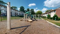 Located in Columbia, Maryland, this outdoor community playground and green space offers a welcoming recreational environment for residents. At its center sits a multi-component play structure featuring a green-roofed platform, dual slides, climbing elements, and overhead monkey bars, all set on a generous wood chip safety surface that cushions the play area. Adjacent seating benches provide a comfortable vantage point for supervising children or simply enjoying the outdoors.

The surrounding landscape is characterized by expansive, well-maintained lawns that roll gently across the grounds, with notable grassy mounds adding natural topographic interest. Mature deciduous trees are distributed throughout the space, offering ample shade and a park-like atmosphere. A network of concrete sidewalks connects various parts of the grounds, providing clear and accessible pathways for pedestrians throughout the community.

Multi-story brick and siding residential buildings frame the perimeter of the playground on multiple sides, creating an enclosed, community-centered feel. Manicured shrubs and greenery line building entrances, contributing to the overall tidiness of the grounds. Under open skies and surrounded by lush foliage, this playground and green space serves as a central outdoor hub for the Columbia, MD community.