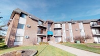 This residential unit is part of a classic multi-story brick building, identifiable by its distinctive teal canopied entrance at address 5365. The exterior presents a solid, traditional architectural character with balconies on upper floors, large windows, and well-kept green grounds that add to its curb appeal.

Inside, the single-floor unit offers a thoughtfully arranged living space. The living room serves as the heart of the home, featuring wall-to-wall carpeting, neutral-toned walls, and a sliding glass door that opens to a private balcony with outdoor views of the surrounding greenery — allowing natural light to fill the room. A wall-mounted air conditioning unit provides in-unit climate control.

Adjacent to the living room is a dedicated dining area, accented by a chandelier overhead and tastefully decorated with wall art, offering an intimate setting for meals.

The kitchen is efficiently designed in a galley layout, featuring warm oak cabinetry, white countertops, a double-basin sink, and white appliances including a refrigerator, dishwasher, and range. Tile flooring and recessed overhead lighting complete this functional space.

Overall, the unit presents a clean, practical, and comfortable living environment with a cohesive neutral palette throughout, making it a versatile and move-in ready space.