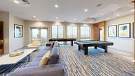 Located at 1855 Saint Francis Street in Reston, VA, this well-appointed amenity lounge offers a generous, open layout across a single floor. The space is defined by an abundance of natural light streaming through large floor-to-ceiling windows and glass doors, framing sweeping panoramic views of the Reston skyline and beyond. A signature blue and cream patterned carpet anchors the main living areas, lending a modern, cohesive aesthetic throughout.

The lounge is thoughtfully divided into distinct functional zones. A relaxed seating area centers around a curved, circular sofa and a wall-mounted flat-screen television above a sleek linear fireplace, creating an inviting focal point for gathering. Adjacent to this is an intimate reading or conversation nook with accent chairs, pendant lighting, and access to an outdoor terrace. Warm wood-paneled accents and built-in shelving add texture and character to the transitions between spaces.

The game room is well-equipped with a full-size billiards table and a shuffleboard table, complemented by casual seating and direct access to the terrace through glass doors. A curved dining area features high-top tables and bar seating set against a sweeping bank of windows, ideal for casual dining or socializing.

The compact kitchenette is finished with marble countertops, a stainless steel sink, and built-in appliances, providing practical support for events and gatherings. A connecting hallway ties the entire space together seamlessly.