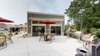 This single-story amenity building is designed to serve as a social hub, combining indoor and outdoor living in a practical and welcoming layout. The exterior is clad in sage green horizontal siding accented by stone veneer detailing at the base, giving the structure a clean, contemporary curb appeal. A white pergola crowns the roofline above a wall of large sliding glass doors, which seamlessly connect the interior to the expansive concrete patio outside. The outdoor patio is generously sized and well-appointed, featuring a dining table with seating and a market umbrella for shade, creating an inviting space for casual gatherings. To one side, a dedicated grilling station equipped with multiple gas grills makes the area ideal for outdoor cooking and entertaining. The sliding doors allow abundant natural light to flow into the interior while maintaining an open, airy connection to the outdoors. A perimeter fence encloses the patio area, offering a sense of privacy while mature trees in the surrounding landscape provide a natural backdrop. The overall design of the building prioritizes functionality and community engagement, making it a versatile space suited for social events and everyday recreational use.