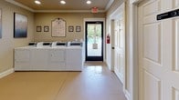 Located at Piney Hills Village in Piney Flats, Tennessee, this single-floor community laundry facility is designed to serve residents with practicality and comfort in mind. The main laundry area is finished with warm beige walls accented by crown molding and recessed lighting, creating a clean and welcoming atmosphere. A row of commercial-grade top-load washers and dryers lines one wall, providing ample capacity for community use. Posted laundry rules and decorative framed artwork add a polished, organized character to the space. A glass-panel door opens directly to the outdoor pool area, allowing natural light to fill the room and offering convenient access between amenities. Adjacent to the laundry area, a separate hallway leads to two clearly marked restrooms — one for each gender — equipped with standard fixtures and fire safety equipment including a mounted fire extinguisher. A small waiting or folding area features a rustic wood-top table with metal legs beneath a decorative round mirror, providing a functional and stylish spot for residents. The smooth flooring throughout, neutral color palette, and thoughtful layout make this facility both easy to navigate and pleasant to use.