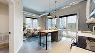 This 2-bedroom, 1-bathroom unit showcases a contemporary industrial aesthetic throughout its single floor. Polished concrete floors flow continuously through the space, complemented by exposed concrete structural columns and visible overhead ductwork that lend an urban loft character. The open-concept kitchen and living area serves as the heart of the unit, featuring a well-appointed kitchen with two-tone cabinetry in white and slate blue, white subway tile backsplash, stainless steel appliances, and a central island with bar seating. Pendant lighting and large windows invite abundant natural light, offering views of the surrounding urban streetscape. The living area provides a comfortable, well-lit retreat anchored by floor-to-ceiling windows. A clean, well-lit hallway lined with polished concrete flooring connects the living spaces to the two bedrooms, with bold modern artwork adding character along the way. The primary bedroom is a well-proportioned room featuring a large window with exterior views. The shared bathroom includes a walk-in shower and clean, minimalist finishes consistent with the rest of the unit. A dedicated laundry area with open shelving provides added convenience. Throughout, the design balances industrial elements with warm, modern touches.