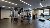 This modern fitness center is thoughtfully designed to support a wide range of workout routines across distinct functional zones. The main exercise floor is finished with durable rubber flooring throughout, providing both safety and comfort underfoot. The strength training area is anchored by a multi-station cable machine system by Life Fitness, complemented by a Synrgy functional training rig, medicine balls, resistance bands, and a variety of accessories for free-form exercise. A dedicated cardio zone lines the perimeter near large windows, housing multiple treadmills and elliptical machines that allow users to exercise while taking in views of the surrounding streetscape and cityscape. Full-length mirrors along the walls visually expand the space and assist with form and technique. A separate studio room features warm hardwood flooring, floor-to-ceiling mirrors, and an open layout ideal for yoga, stretching, group fitness, or personal training sessions. The entryway to the facility includes a built-in cubby storage unit, a water fountain station, and a wall-mounted digital room management panel, ensuring a streamlined experience for users. Linear LED lighting throughout provides consistent, bright illumination across all areas, contributing to the clean, contemporary aesthetic of the overall facility.