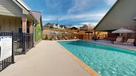 Located at 101 Fox Grape Loop in Weaverville, North Carolina, this outdoor amenity area serves as the community pool facility for Encore at Lake James. The centerpiece is a generously sized rectangular swimming pool with crystal-clear blue water, surrounded by a wide concrete sundeck designed to accommodate ample lounge seating and umbrellas for shade. A decorative living green wall prominently displays the Encore community branding, serving as a striking visual focal point adjacent to the pool house structure. The pool house features a pitched roof with a covered pavilion area, providing a sheltered gathering space for residents. An accessibility lift is installed at the pool's edge, ensuring inclusive access for all users. The pool area is fully enclosed by black metal fencing with a gated entry, and pool rules and safety signage are clearly posted at the entrance. Additional outdoor seating is available under a wooden pergola on the far side of the deck, offering a shaded retreat. The surrounding landscape reveals the rolling hillside community of homes, with open sky views that lend a bright, airy atmosphere. The overall design balances functionality and aesthetic appeal, making this pool area a welcoming and well-appointed outdoor gathering space for the community.