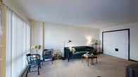 This single-floor residence offers a thoughtfully arranged living space with a calm, understated aesthetic. The main living area is centered around plush wall-to-wall carpeting that adds warmth and comfort underfoot, complemented by smooth, light-colored walls that create a bright and airy atmosphere. The layout is open and practical, providing ample room for seating and relaxed social gatherings. A tufted dark green sofa serves as the focal point of the living area, paired with wooden accent tables and a sleek metal-framed floor lamp that adds both function and style. Decorative touches such as framed artwork leaning against the walls, a small shelving unit, and a vase of dried flowers contribute a curated, lived-in character to the space. A dining or reading nook is visible near the corner, furnished with classic wooden chairs, adding a versatile area for multiple uses. The neutral ceiling and walls throughout maintain a cohesive, clean visual flow from room to room. Natural and ambient lighting work together to keep the interiors feeling open and welcoming. A doorway visible in the background suggests additional rooms beyond the main living space, hinting at a well-connected and functional single-floor floor plan overall.