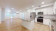 This single-floor unit presents a clean, contemporary design with a cohesive neutral color palette throughout. The open-concept kitchen and living area serves as the central hub of the space, anchored by a large center island with a dark quartz countertop that provides ample workspace and seating. The kitchen is well-appointed with white shaker-style cabinetry, a white subway tile backsplash, stainless steel appliances including a French door refrigerator with ice and water dispenser, an electric range, and an over-the-range microwave. Light wood-look flooring extends throughout the kitchen and living area, contributing to the bright, airy atmosphere enhanced by recessed lighting. The spacious living area offers generous room for various furniture arrangements and receives natural light through windows near the entry.

The bedrooms feature neutral wall-to-wall carpeting, white walls, and window treatments for privacy, with at least one bedroom equipped with a ceiling fan. Multiple windows in select bedrooms allow for abundant natural light.

The bathrooms are finished with white shaker cabinetry, taupe quartz countertops, black hardware accents, and large mirrors. The primary bathroom includes a dual-sink vanity and a glass-enclosed shower. A dedicated in-unit laundry area houses a full-size Whirlpool washer and dryer.