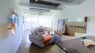 This single-floor space showcases a sleek, modern design centered around an open-plan living area that seamlessly integrates multiple functional zones. The polished concrete flooring runs throughout, lending an industrial-chic character while reflecting the abundant natural light that pours in through expansive floor-to-ceiling windows along one wall. These large glazed panels frame outdoor views and create a bright, airy atmosphere.

The living area is anchored by a plush, oversized sectional sofa in soft grey, complemented by layered textiles including patterned cushions and a warm orange throw. A circular coffee table sits atop a deep blue area rug, adding contrast to the neutral palette. A wall-mounted entertainment unit in warm wood tones provides storage and display space, while a large-format artwork adds a bold focal point.

Structural ceiling details, including recessed paneling and exposed mechanical elements, add architectural interest overhead. Track lighting supplements the natural light. A dining area with a light wood table is visible toward the windows, and dark granite countertops at the foreground hint at a well-appointed kitchen nearby. The overall ambiance is polished, open, and contemporary.