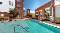 This outdoor pool area is set within a well-designed courtyard in Baton Rouge, Louisiana, framed by multi-story brick and contemporary-clad residential buildings that create a sense of enclosure and privacy. The pool itself is expansive, featuring clear turquoise water with a geometric layout that includes both shallow entry points and deeper sections, all accessible via stainless steel handrails. The surrounding pool deck is finished in smooth concrete, providing ample space for lounge chairs and relaxed sunbathing under the open Louisiana sky.

Lush landscaping lines the perimeter, with tropical plantings including palm trees and ornamental ground cover that reinforce the warm, Southern atmosphere. A pergola-covered outdoor lounge and grilling station is positioned at one end of the courtyard, furnished with seating and outdoor cooking equipment, making it well-suited for social gatherings. The brick and stone exterior detailing of the adjacent building walls adds architectural texture and visual warmth to the space. Large glass doors provide interior access directly to the pool deck, enhancing the connection between indoor and outdoor areas. Overall, the space offers an inviting, resort-style outdoor environment ideal for recreation and relaxation.