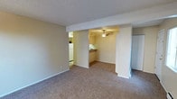 This single-floor residential unit offers a practical and straightforward layout designed for comfortable everyday living. The main living area is finished with wall-to-wall carpeting in a neutral tone, contributing to a warm and understated ambiance throughout. Textured ceilings add a classic touch to the interior. The open floor plan connects the living space to a secondary area, likely a dining or secondary sitting room, through a wide archway framed by a structural beam. This adjoining space features a ceiling fan, providing both airflow and a functional lighting element. A breakfast bar serves as a natural transition point between the living areas and the kitchen, offering casual seating and a sense of openness. The kitchen is equipped with cabinetry in a dark finish and includes a white refrigerator, providing practical storage and essential appliances. Neutral-toned walls throughout keep the space feeling clean and versatile. A closet is visible along one wall of the living area, offering additional built-in storage. The overall aesthetic is simple and functional, making the space well-suited for those seeking a move-in-ready, low-maintenance living environment.