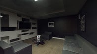 This single-floor space is designed with a distinctly modern and moody aesthetic, combining dark textured wall finishes with carefully curated furnishings to create an intimate, lounge-like environment. The walls throughout are clad in a deep charcoal grasscloth-style texture, lending depth and sophistication to the interior while establishing a cohesive, dramatic tone across the room.

A standout architectural feature is the expansive built-in media wall, constructed in crisp white and composed of an arrangement of open shelving, closed cabinetry, and drawers of varying sizes. A large flat-screen television is centrally mounted within this unit, which also accommodates audio-visual components. The contrast between the white media wall and the surrounding dark walls creates a striking visual focal point.

Seating consists of contemporary gray upholstered sofas and accent chairs arranged around a sculptural glass-top coffee table with a geometric metallic base. The flooring features a neutral-toned carpet that softens the overall palette. Recessed ceiling panels provide overhead illumination, supplemented by flush lighting that highlights the room's structured layout.

Black-and-white framed artwork decorates the walls, adding a curated touch. A standard access door with an illuminated exit sign is positioned along one wall, indicating this space is part of a larger commercial or multi-use building.