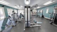 This fitness center offers a comprehensive workout environment designed to accommodate a wide range of exercise needs. The space is finished with teal-painted walls and durable rubber flooring, creating a clean, energetic atmosphere conducive to physical activity. Bright flush-mounted lighting panels combine with natural light from multiple windows and French doors to ensure the room remains well-lit throughout the day. Ceiling fans provide additional air circulation, enhancing comfort during workouts.

The equipment layout is thoughtfully organized, with a prominent Life Fitness cable/weight stack machine positioned centrally, complemented by an adjustable workout bench nearby. The cardio section features multiple treadmills and at least one recumbent-style machine, positioned toward the windowed wall to allow exercisers to enjoy natural light. On the opposite side, a power rack with weight plates and a barbell provides space for strength training. Additional selectorized machines are arranged throughout, offering options for targeted muscle group training. The overall layout balances open floor space with a diverse selection of professional-grade equipment, making this fitness center suitable for both cardio and strength-focused training routines.
