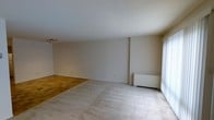 This single-floor space presents an open layout that seamlessly connects the living and dining areas. The flooring transitions distinctly between the two zones, with warm parquet hardwood defining the dining area and light-toned carpet covering the living space, creating a natural visual division without the need for walls or partitions. The neutral beige walls provide a clean, versatile backdrop throughout, lending the space a calm and understated atmosphere.

A textured popcorn ceiling runs across the entirety of the space, a characteristic finish consistent with the building's construction era. A track lighting fixture is mounted near the dining area, offering directed illumination for that zone. On the opposite end of the room, vertical blinds frame a large window or sliding door, allowing for ample natural light to filter into the living area and offering the flexibility to control privacy and brightness. A baseboard-style radiator unit is positioned beneath the window, indicating a radiator-based heating system. The overall ambiance of the space is bright, neutral, and move-in ready, offering a solid foundation for a variety of interior design styles.