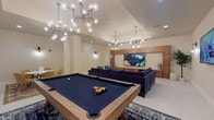 This single-floor amenity lounge is designed as a multifunctional social space, combining entertainment, relaxation, and casual gathering areas within a cohesive, contemporary aesthetic. The layout is thoughtfully zoned to accommodate a variety of activities simultaneously. A full-size pool table with navy felt serves as the centerpiece of the gaming area, positioned beneath striking sputnik-style chandeliers with globe bulbs that illuminate the space with a warm, ambient glow.

The lounge area features a large, deep-blue velvet sectional sofa anchored by a live-edge wood coffee table atop a patterned area rug, creating an inviting focal point. A floor-to-ceiling wood-paneled media wall houses a large flat-screen television, complemented by additional wall-mounted screens for flexible viewing. Built-in cabinetry with decorative shelving adds both storage and visual interest to this area.

A separate seating nook with two mid-century modern blue accent chairs is paired with a warm-toned wood built-in unit featuring recessed display shelving. A casual dining alcove accommodates a round white table surrounded by mustard-yellow upholstered chairs atop a traditional area rug. Throughout, light tile flooring, cream-toned walls, and sleek wall sconces maintain a clean, polished atmosphere.