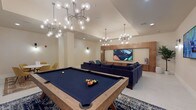 This single-floor amenity lounge is designed as a multifunctional social space, combining entertainment, relaxation, and casual gathering areas within a cohesive, contemporary aesthetic. The layout is thoughtfully zoned to accommodate a variety of activities simultaneously. A full-size pool table with navy felt serves as the centerpiece of the gaming area, positioned beneath striking sputnik-style chandeliers with globe bulbs that illuminate the space with a warm, ambient glow.

The lounge area features a large, deep-blue velvet sectional sofa anchored by a live-edge wood coffee table atop a patterned area rug, creating an inviting focal point. A floor-to-ceiling wood-paneled media wall houses a large flat-screen television, complemented by additional wall-mounted screens for flexible viewing. Built-in cabinetry with decorative shelving adds both storage and visual interest to this area.

A separate seating nook with two mid-century modern blue accent chairs is paired with a warm-toned wood built-in unit featuring recessed display shelving. A casual dining alcove accommodates a round white table surrounded by mustard-yellow upholstered chairs atop a traditional area rug. Throughout, light tile flooring, cream-toned walls, and sleek wall sconces maintain a clean, polished atmosphere.