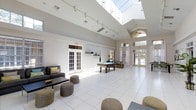 This single-story recreational space is defined by its dramatic, soaring ceilings that create an expansive and airy atmosphere throughout. A large skylight positioned at the apex of the ceiling floods the interior with natural light, complementing the bright white walls and clean white tile flooring that extend across the entire space.

The layout is open and versatile, accommodating multiple leisure zones. A lounge area near the entrance features a dark sectional sofa accented with colorful throw pillows, paired with a low coffee table and woven pouf seating. The central area is anchored by a full-size billiards table, serving as a natural focal point for social activity.

Tall French doors line multiple walls, offering direct access to the outdoors and drawing in additional natural light while providing views of surrounding greenery. A ceiling fan is mounted near one of the windowed sections, enhancing ventilation and comfort. Track lighting installed along the ceiling supplements natural light during evening hours.

The overall design is clean, modern, and functional, with a neutral palette that creates an inviting, communal environment well-suited for recreation and social gatherings.